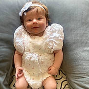 Nevaeh is registered to the contest to win money with this photo: baby, smiling, white_dress, headband, puffed_sleeves, infant, cushion, pillow, soft_fabric, sitting, happy, indoors, cute, child, portrait, legs, hands, pattern, cozy, home