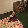 baby, infant, child, play_mat, plush_toy, toy, carpet, indoor, heater, wall, outlet, plaid_dress, dress, sitting, floor, hand_in_mouth, cute, portrait, colorful, green_carpet