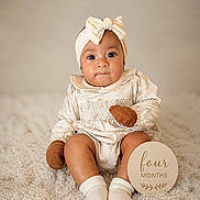 Ivy is registered to the contest to win money with this photo: baby, infant, four_months, headband, bow, mittens, socks, cream_outfit, carpet, wooden_sign, portrait, cute, child, sitting, indoors, soft_lighting, neutral_background, smile, young_child, cozy