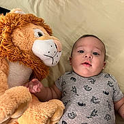 Alan is registered to the contest to win money with this photo: baby, infant, plush_toy, lion, stuffed_animal, bed, bedding, clothing, onesie, child, cute, soft, toy, indoor, portrait, lying_down, person, young_child, face, expression