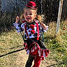 child, clown_costume, swing, playground, grass, sunlight, hat, face_paint, boots, red, black, white, striped_skirt, pigtails, outdoor, fence, chain, happy, tongue_out, smiling
