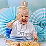baby, birthday, cake, celebration, child, crown, blue_balloons, paper_fans, smiling, messy, wooden_spoon, sitting, white_shirt, shorts, party, first_birthday, happy, portrait, indoor, gold_chain