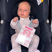 Ocean is registered to the contest to win money with this photo: baby, infant, car_seat, child, straps, pink_clothing, sign, wide_eyes, surprised_expression, sitting, indoor, safety, person, newborn, cute, portrait, head, feet, holding, closeup