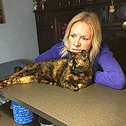 Ségolène Frechin a rejoint le concours — aidez-le/la à gagner de superbes lots ! woman, cat, tortoiseshell_cat, table, purple_sweater, furniture, indoor, relaxed, decorations, wooden_cabinet, pet, companion, looking_away, resting, home, cozy, person, animal, domestic, curiosity