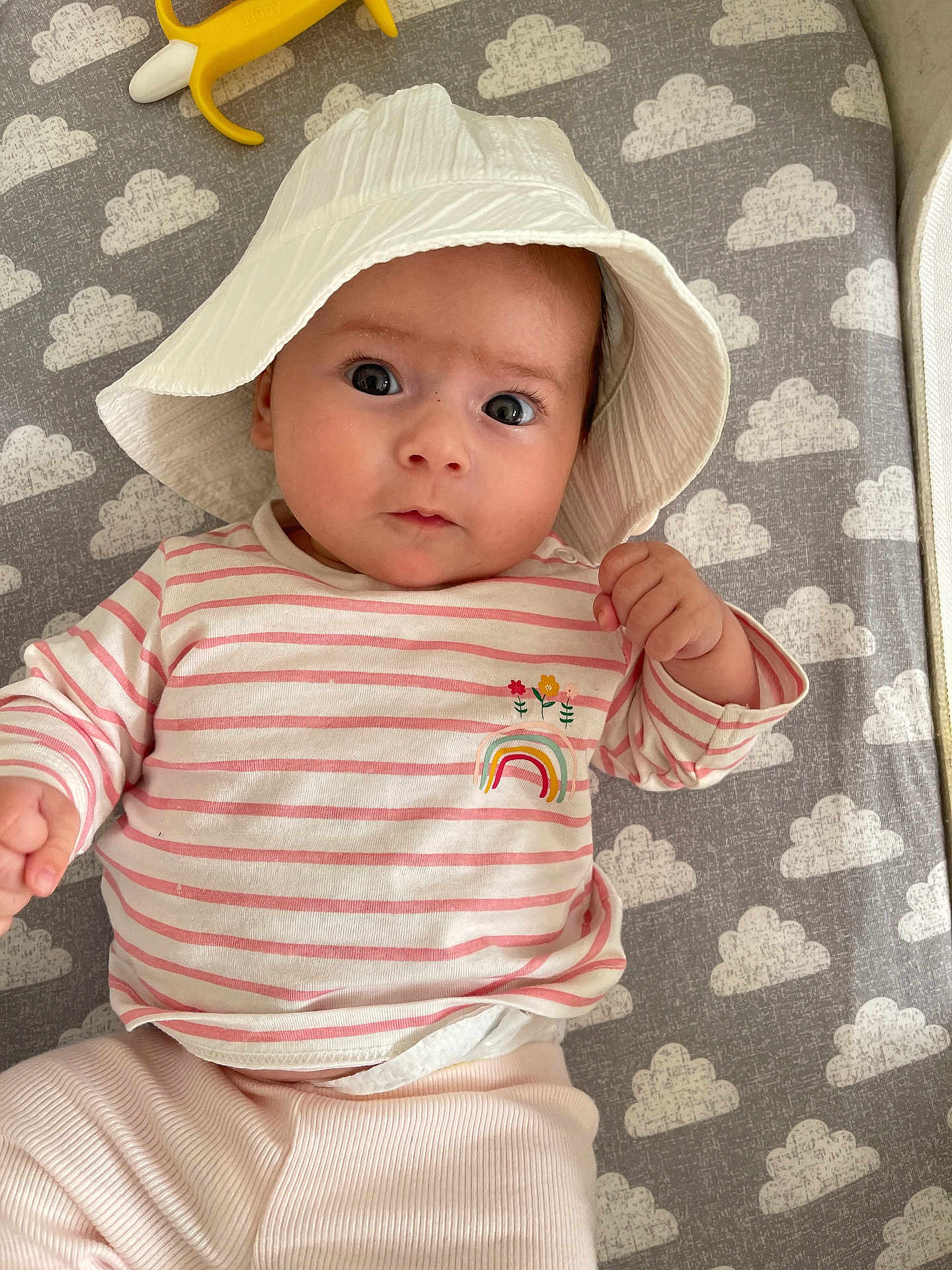 Amber is registered to the contest to win money with this photo: baby, infant, hat, white_hat, striped_shirt, rainbow_patch, pink_pants, cloud_pattern, mat, lying_down, cute, child, indoors, soft_lighting, portrait, person, small_hands, wide_eyes, clothing, baby_toy