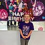 Braylee is registered to the contest to win money with this photo: child, girl, birthday, banner, crown, smile, tshirt, purple_clothing, animated_characters, party, celebration, indoor, standing, happy, casual_clothing, grey_pants, balloon, background, reflection, floor
