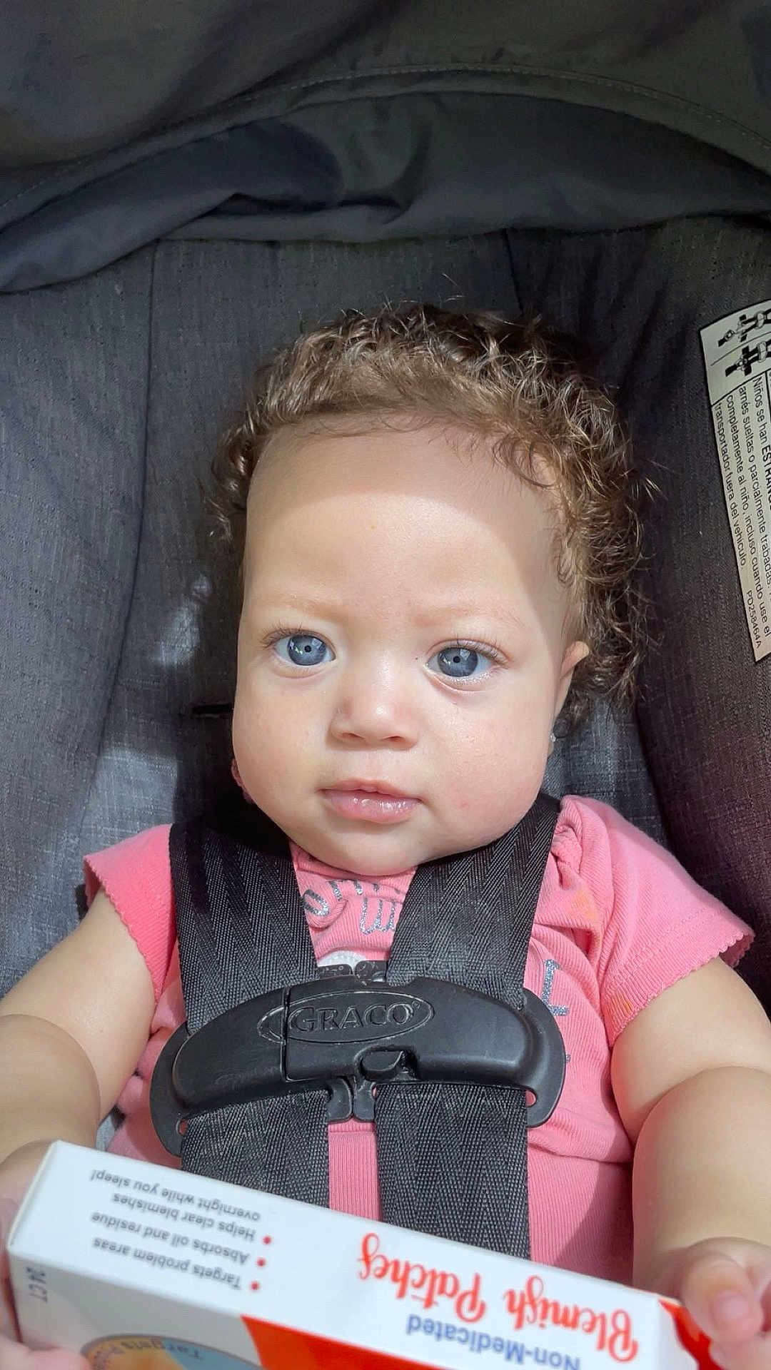 Journi is registered to the contest to win money with this photo: baby, child, car_seat, harness, curly_hair, blue_eyes, pink_shirt, box, benzyl_pads, close_up, indoors, seatbelt, sitting, infant, person, cute, portrait, skin, face, hand