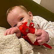 Hazel joined the competition — help win amazing prizes! baby, child, plush_toy, dragon, blanket, cozy, indoors, smiling, cute, hand, face, clothing, person, soft_toy, lying_down, infant, playful, warm_lighting, bedroom, expression