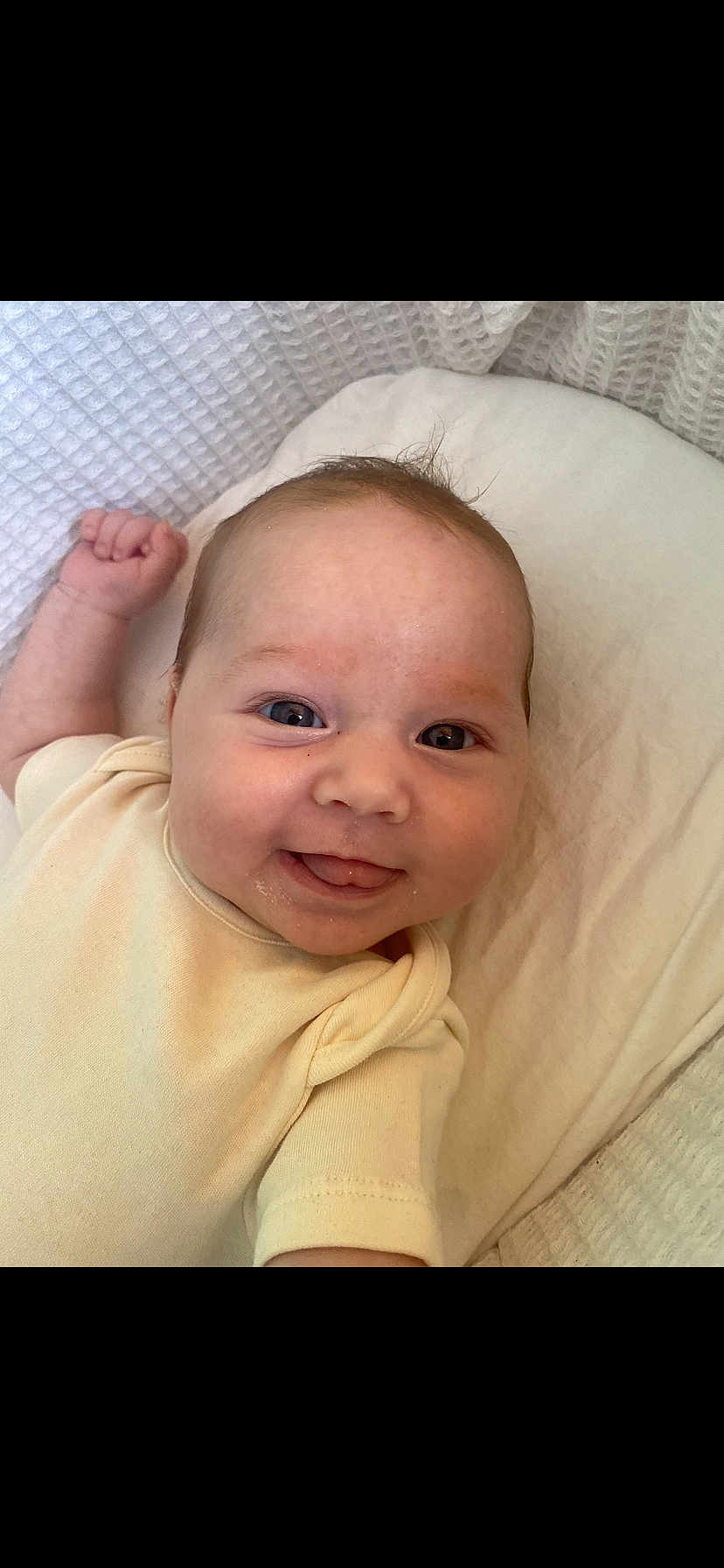 Hazel is registered to the contest to win money with this photo: baby, smiling, tongue_out, onesie, blanket, infant, cute, face, happy, child, skin, head, lying_down, soft_texture, closeup, indoors, person, newborn, expression, cheerful