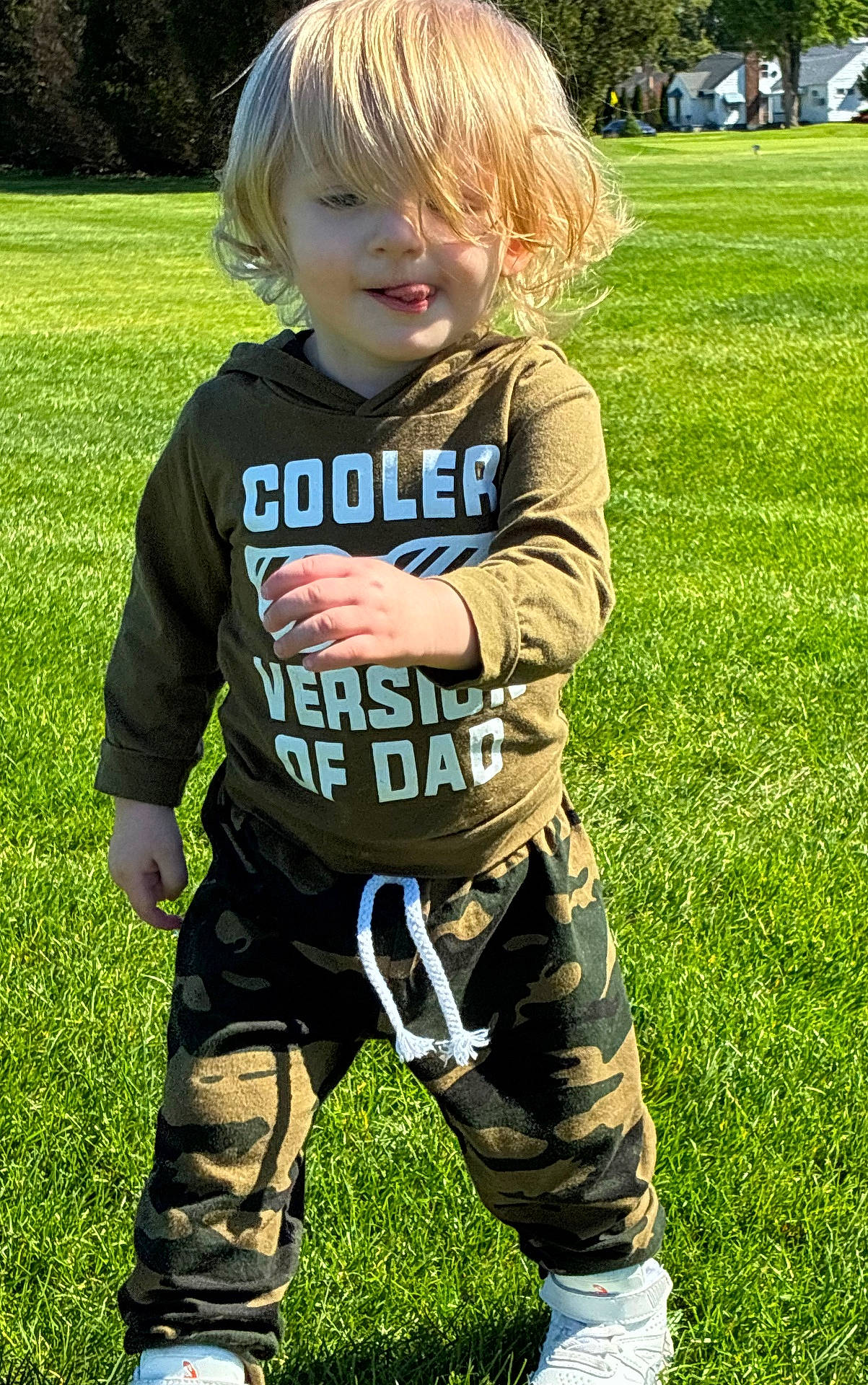 Kolsen is registered to the contest to win money with this photo: blonde_hair, camo_pants, casual_clothing, child, cute, daylight, grass, greenery, happy, hoodie, nature, outdoor, playful, smiling, standing, sunny, toddler, tongue_out, white_shoes, young_child