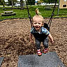 toddler, child, swing, playground, park, grass, trees, picnic_table, shoes, clothing, smiling, happy, hair, topknot, outdoor, fun, woodchips, daylight, person, recreation