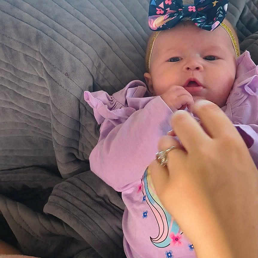 Rylie Miller is registered to the contest to win money with this photo: adult_hand, baby, blanket, blue, bow, clothing, cozy, cute, floral_pattern, hand, headband, indoor, infant, lying_down, pacifier, person, pink, purple, toddler, unicorn