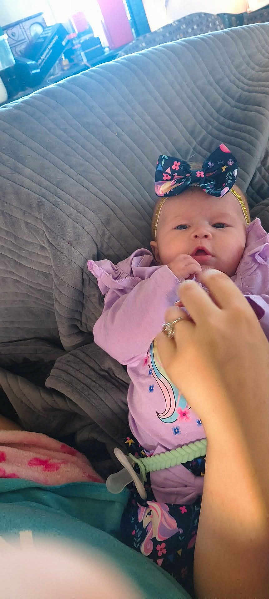 Rylie Miller is registered to the contest to win money with this photo: adult_hand, baby, blanket, blue, bow, clothing, cozy, cute, floral_pattern, hand, headband, indoor, infant, lying_down, pacifier, person, pink, purple, toddler, unicorn