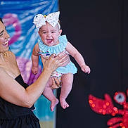 Everleigh joined the competition — help win amazing prizes! baby, blue_outfit, blurred_background, bow, celebration, child, colorful, cute, decoration, happy, headband, holding, indoor, joyful, laughing, party, person, red_crab, smile, woman