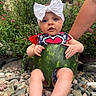baby, bow, child, clothing, curious, cute, flowers, greenery, hand, headband, nature, outdoor, pebbles, plants, portrait, rocks, sitting, summer, support, watermelon