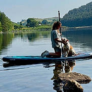 Dora joined the competition — help win amazing prizes! person, dog, paddleboard, lake, water, reflection, outdoor, nature, trees, rocks, hill, greenery, calm, summer, casual_clothing, shorts, sneakers, pet, animal, sunny