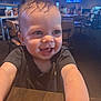 baby, child, smile, happy, face, hair, earring, black_shirt, table, restaurant, chair, background, television, indoor, person, cute, expression, young_child, skin, light
