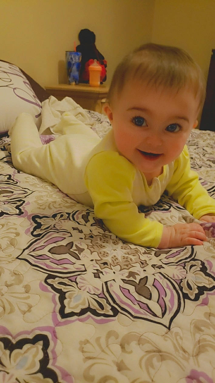 Briana is registered to the contest to win money with this photo: baby, baby_toddler_clothing, bed, cheek, child, comfort, floor, flooring, fun, happy, head, linens, pattern, person, room, sitting, skin, smile, t_shirt, toddler