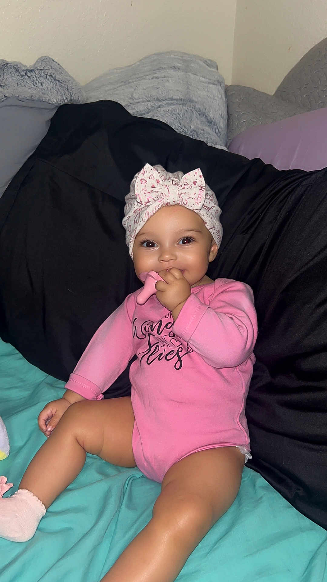 Ivori Jane Blakeley joined the competition — help win amazing prizes! baby, child, pink_onesie, headwrap, teething_toy, smiling, sitting, indoor, pillow, bed, blanket, cute, infant, happy, skin, hand, foot, sock, face, person