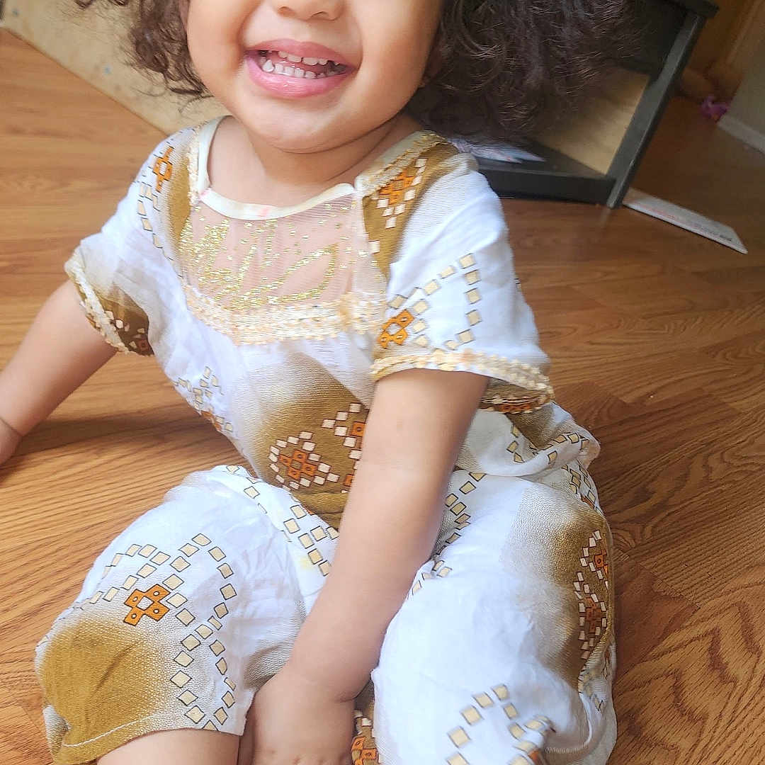 Rabia is registered to the contest to win money with this photo: baby, bright, casual, cheerful, child, curly_hair, cute, foot, hand, happy, indoor, patterned_clothing, person, portrait, sitting, smiling, toddler, white_clothing, wooden_floor, young_child