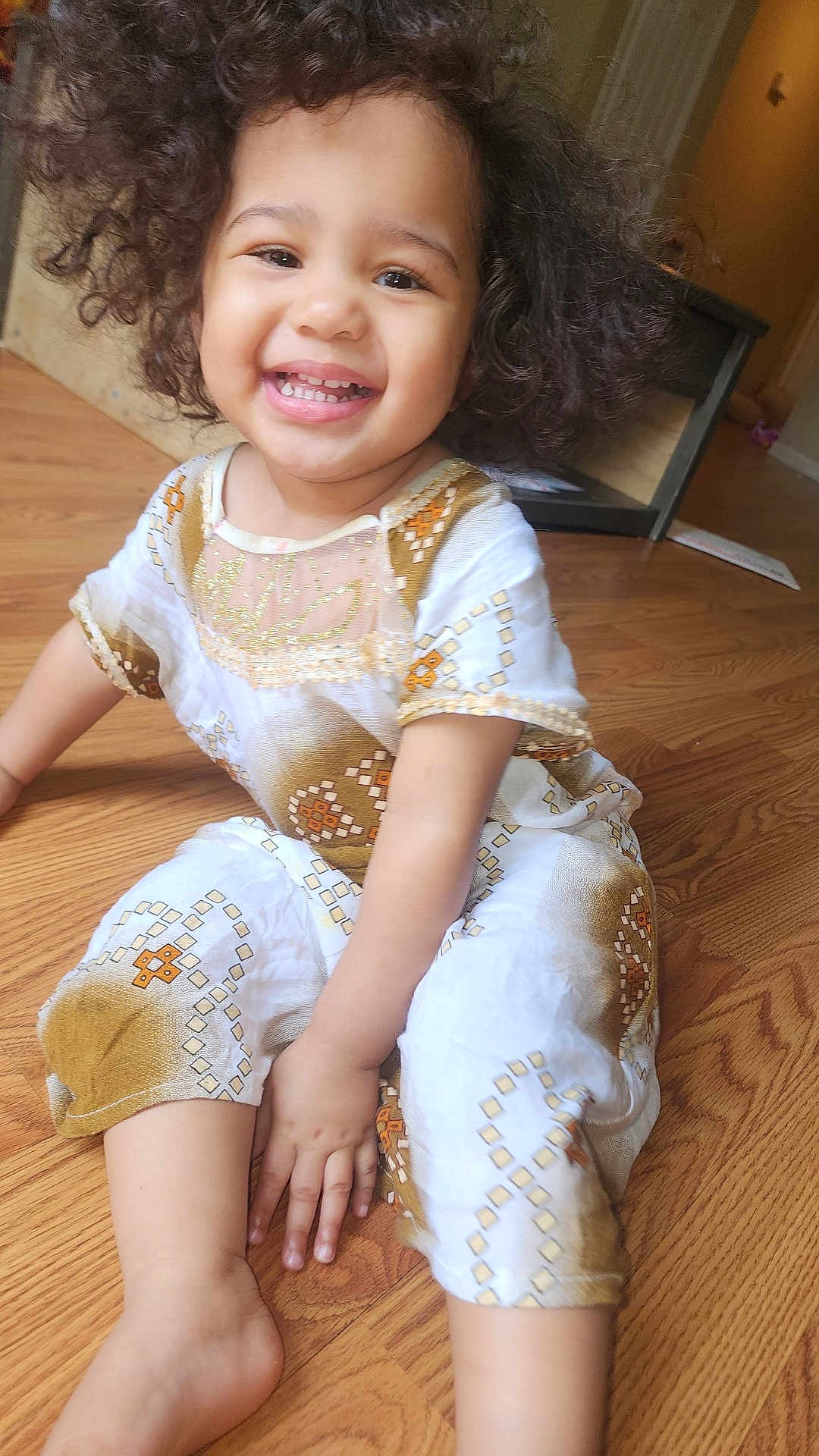 Rabia is registered to the contest to win money with this photo: toddler, child, smiling, curly_hair, indoor, wooden_floor, white_clothing, patterned_clothing, happy, sitting, person, baby, cute, hand, foot, portrait, young_child, cheerful, casual, bright