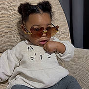 Primrose is registered to the contest to win money with this photo: toddler, child, sunglasses, heart_shaped_glasses, sweater, cat_design, seated, chair, indoors, curly_hair, fashion, cute, cozy, portrait, young_child, casual_clothing, expression, comfortable, home, relaxed