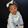 baby, blue_clothing, bow, child, cozy, curious, cute, dark_background, expression, face, indoors, infant, knitted_outfit, person, portrait, sitting, small_hands, soft_lighting, white_bow, young