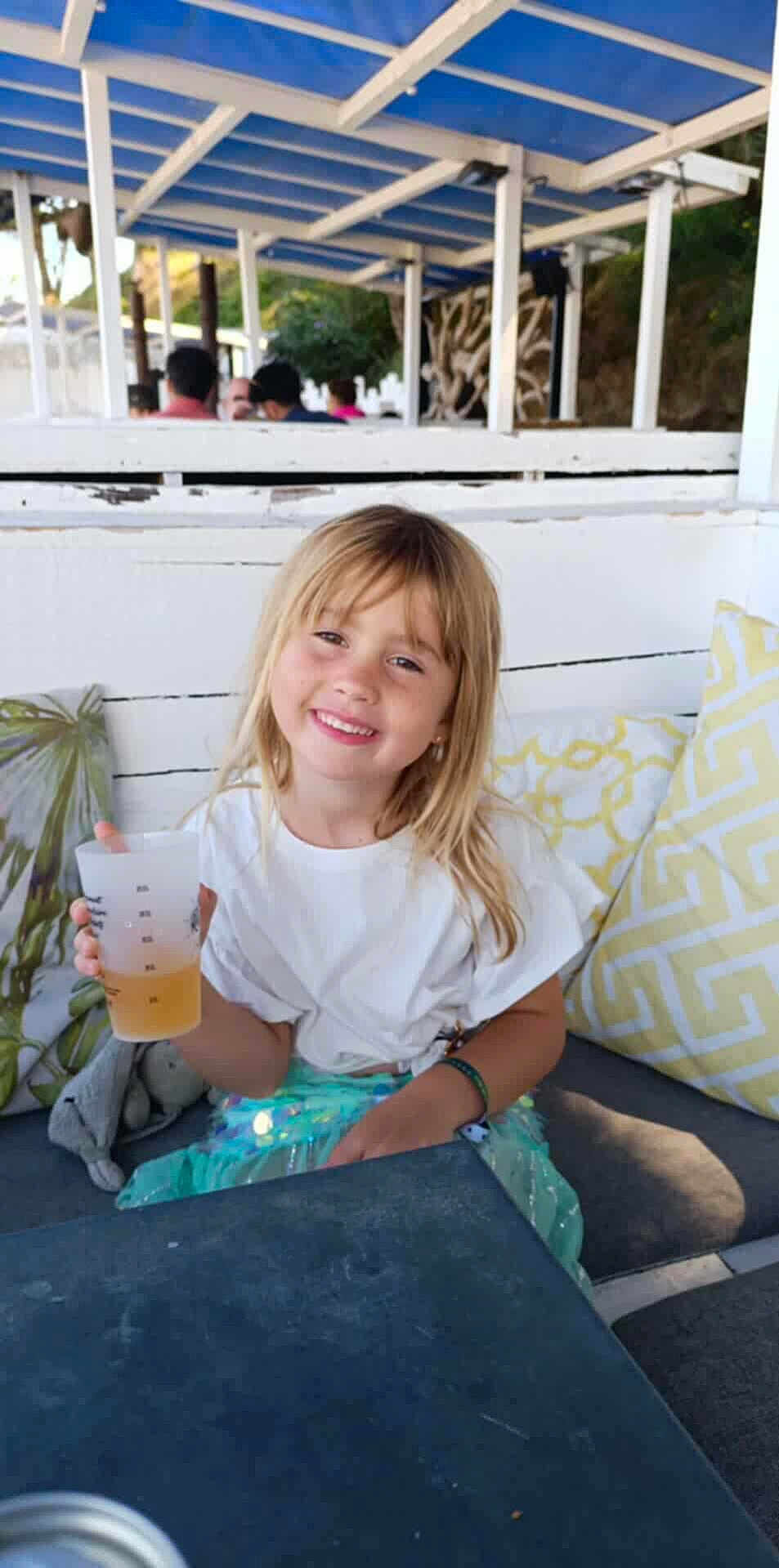 Romy a rejoint le concours — aidez-le/la à gagner de superbes lots ! child, girl, smile, blonde_hair, white_shirt, plastic_cup, drink, cushion, bench, outdoor, canopy, table, happy, casual, daylight, relaxing, seating, people_in_background, bracelet, park