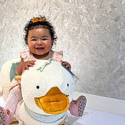 Raven is registered to the contest to win money with this photo: baby, child, smiling, curly_hair, bow, plush_toy, rocking_toy, duck, pink_clothing, socks, happy, indoor, floor, wooden_rocker, cute, portrait, playful, toy, infant, toddler