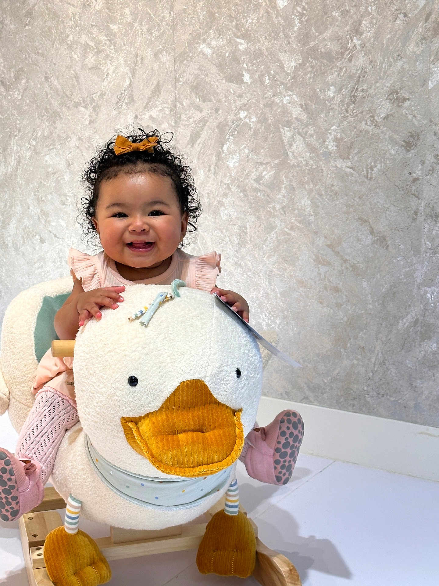 Raven is registered to the contest to win money with this photo: baby, child, smiling, curly_hair, bow, plush_toy, rocking_toy, duck, pink_clothing, socks, happy, indoor, floor, wooden_rocker, cute, portrait, playful, toy, infant, toddler
