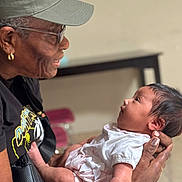 Jasmine joined the competition — help win amazing prizes! elderly_person, baby, holding, smiling, indoor, white_clothing, cap, glasses, earrings, skin, face, hands, portrait, interaction, love, affection, tenderness, people, focus, background_blur