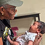 elderly_person, baby, holding, smiling, indoor, white_clothing, cap, glasses, earrings, skin, face, hands, portrait, interaction, love, affection, tenderness, people, focus, background_blur