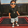 toddler, child, boy, sunglasses, necklace, black_tshirt, denim_shorts, cross_print, sneakers, rug, door, indoors, smiling, standing, fashion, casual, happy, portrait, footwear, clothing