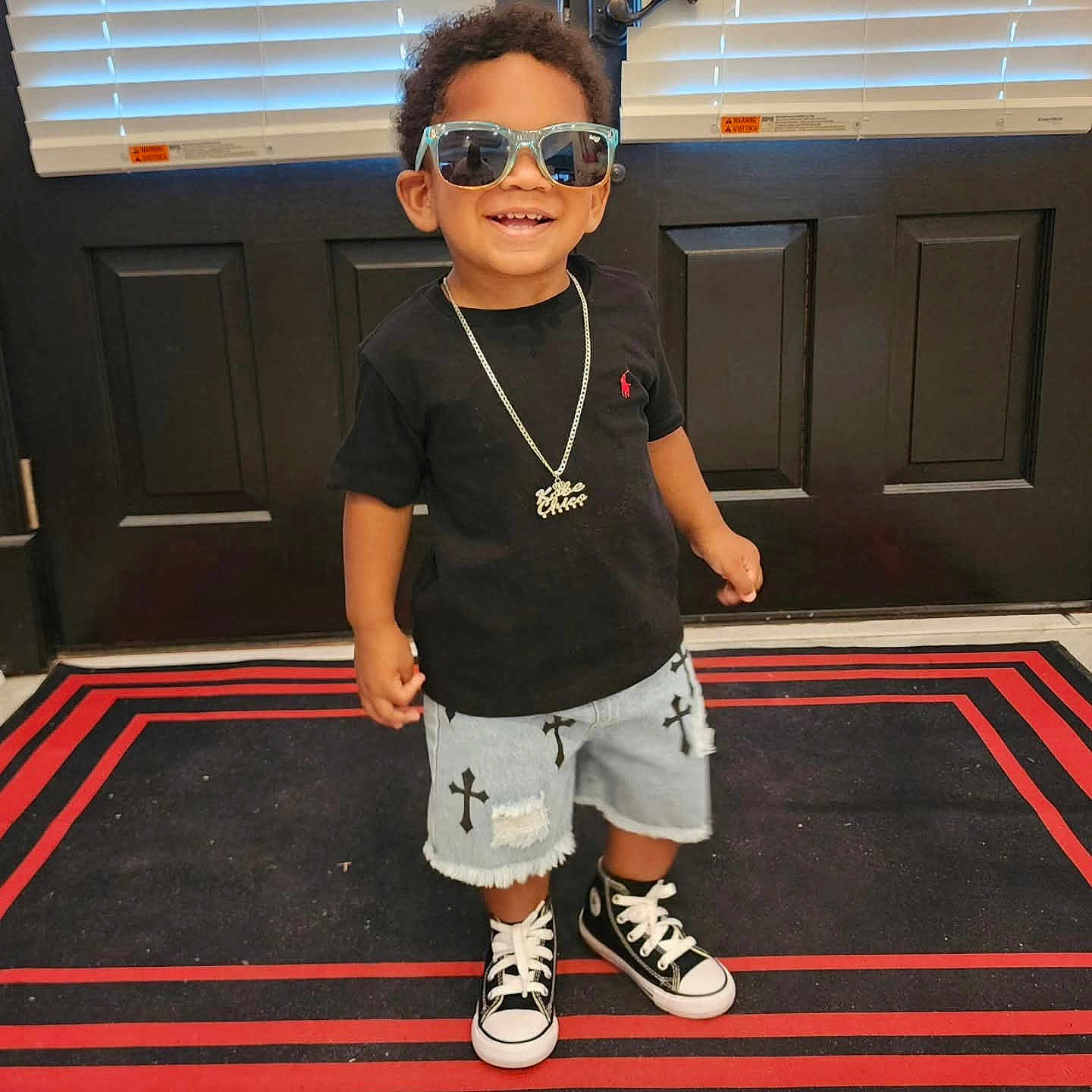 Kobe is registered to the contest to win money with this photo: toddler, child, boy, sunglasses, necklace, black_tshirt, denim_shorts, cross_print, sneakers, rug, door, indoors, smiling, standing, fashion, casual, happy, portrait, footwear, clothing