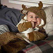 Lila joined the competition — help win amazing prizes! baby, child, onesie, hood, antlers, bed, blanket, plaid, hand, smile, indoor, cozy, cute, infant, face, clothing, young, person, lying_down, warm
