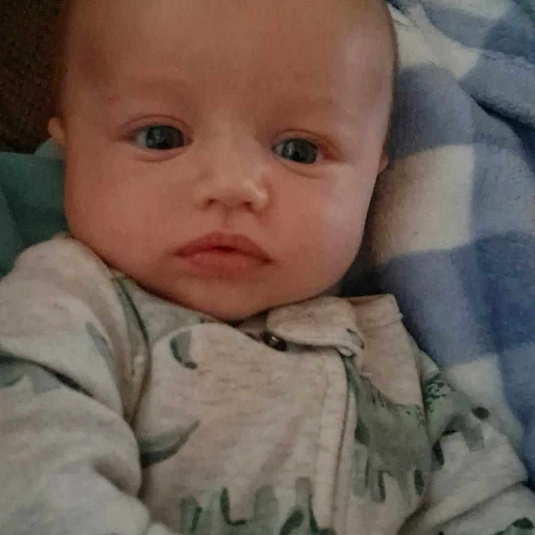 Logan is registered to the contest to win money with this photo: baby, infant, child, face, portrait, wide_eyes, calm, lying_down, blanket, blue_blanket, onesie, clothing, couch, indoor, home, close_up, head, eyes, baby_clothing, photography