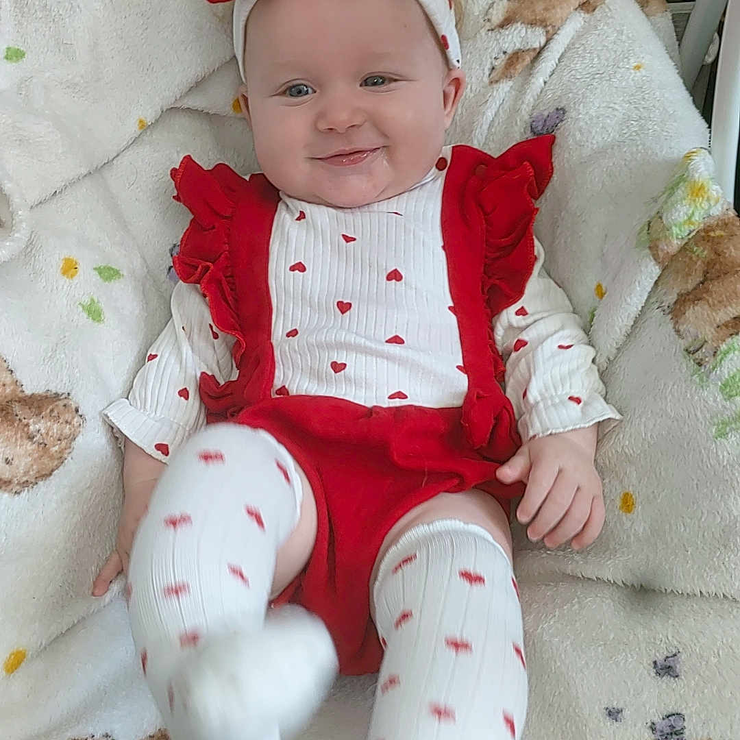 Skylar-Rose is registered to the contest to win money with this photo: baby, blanket, butterfly_pattern, child, cute, footwear, happy, headband, heart_pattern, indoor, infant, person, playful, portrait, red_clothing, sitting, smile, soft_texture, teddy_bear_pattern, white_clothing