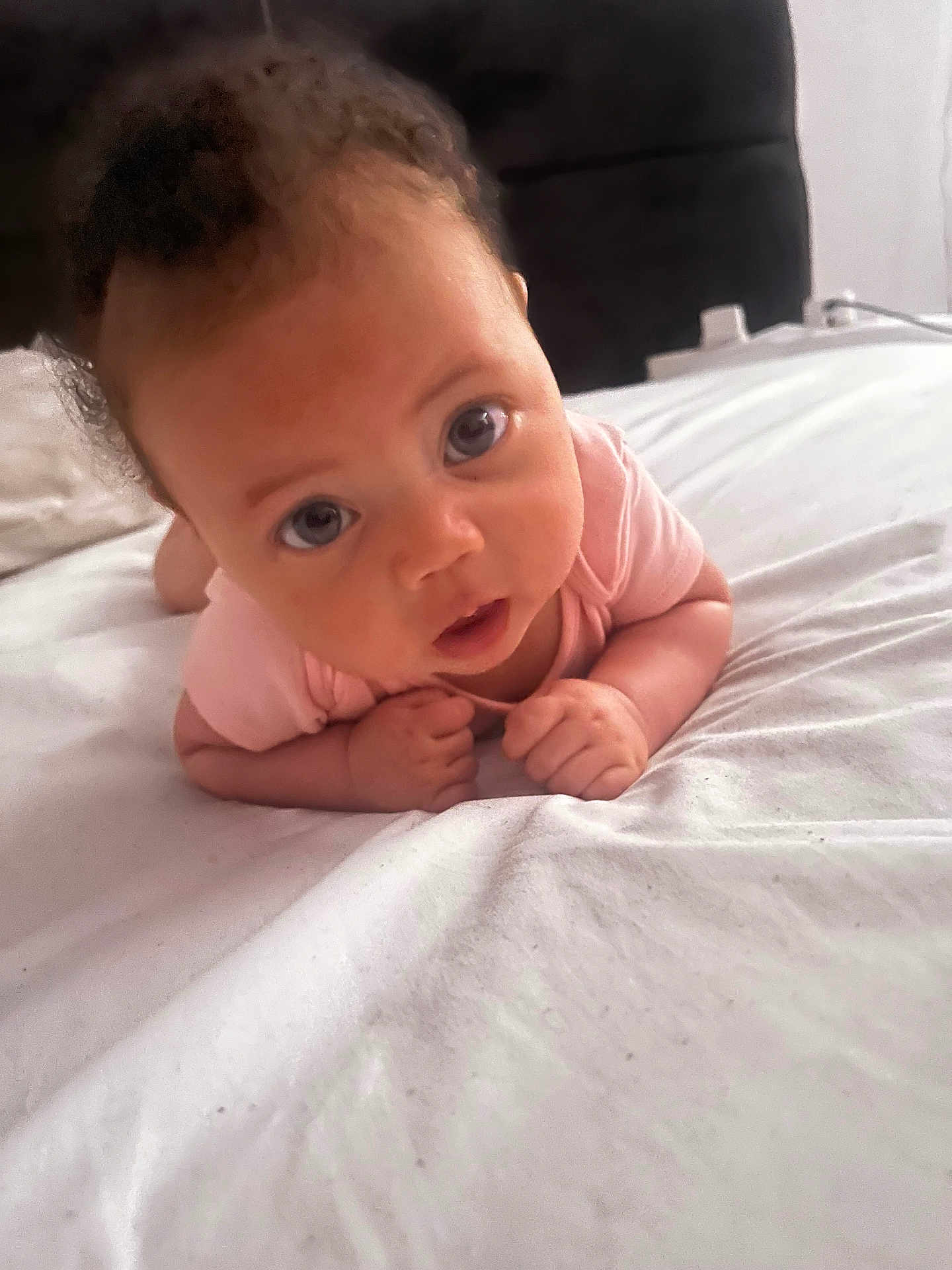 Azriah is registered to the contest to win money with this photo: baby, infant, tummy_time, pink_onesie, bed, white_bedspread, face, eyes, hands, curious, portrait, indoor, child, young, skin, head, expression, person, candid, resting