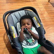 Asante is registered to the contest to win money with this photo: baby, child, car_seat, remote_control, floor, wooden_floor, clothing, green_shorts, white_shirt, socks, indoor, person, face, hands, cute, infant, seated, teething, black_hair, expression