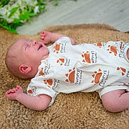 Vinnie is registered to the contest to win money with this photo: baby, newborn, infant, sleeping, smiling, onesie, clothing, blanket, brown, texture, floor, wooden_floor, greenery, plant, peaceful, cute, small, hands, feet, indoors