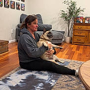 Taco is registered to the contest to win money with this photo: casual_clothing, couch, dog, dresser, furniture, holding, home_decor, indoor, living_room, pet, photographs, plant, pug, relaxed, rug, sitting, wall, woman, wooden_floor, yoga_mat