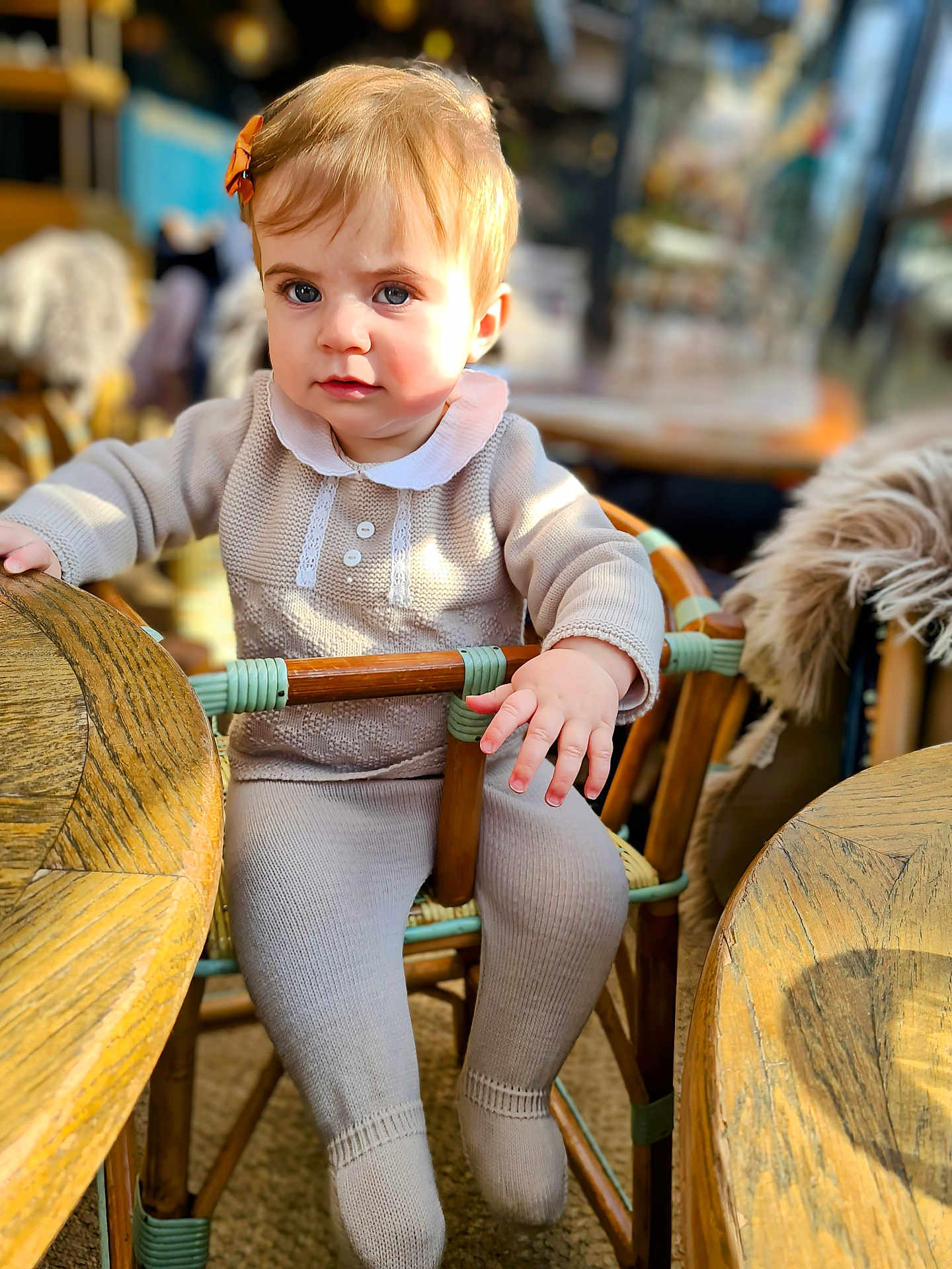 Mia participe au concours pour gagner de l'argent avec cette photo : toddler, child, baby, sitting, chair, wooden_furniture, bow, hair_accessory, indoors, table, knitwear, beige_clothing, cute, portrait, person, young_child, sunlight, cozy, cafe, expression