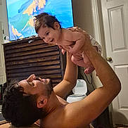 Cassiel is registered to the contest to win money with this photo: baby, infant, parent, father, family, smiling, laughing, playful, indoors, bedroom, television, furniture, bed, blanket, door, lifting, bonding, portrait, home, child