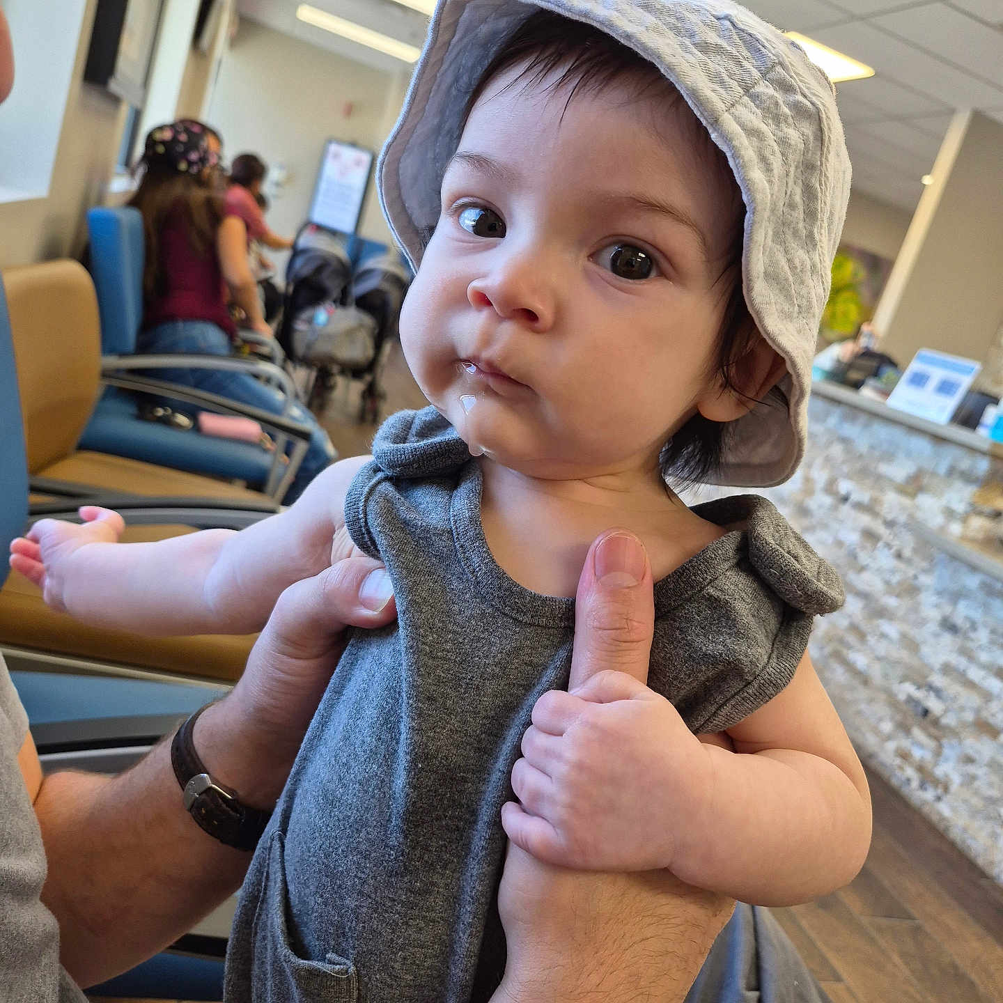 Cassiel is registered to the contest to win money with this photo: baby, child, infant, portrait, wide_eyes, sun_hat, hat, gray_clothing, sitting, held, adult, hands, airport, waiting_area, chairs, indoor, background, floor, counter, photography