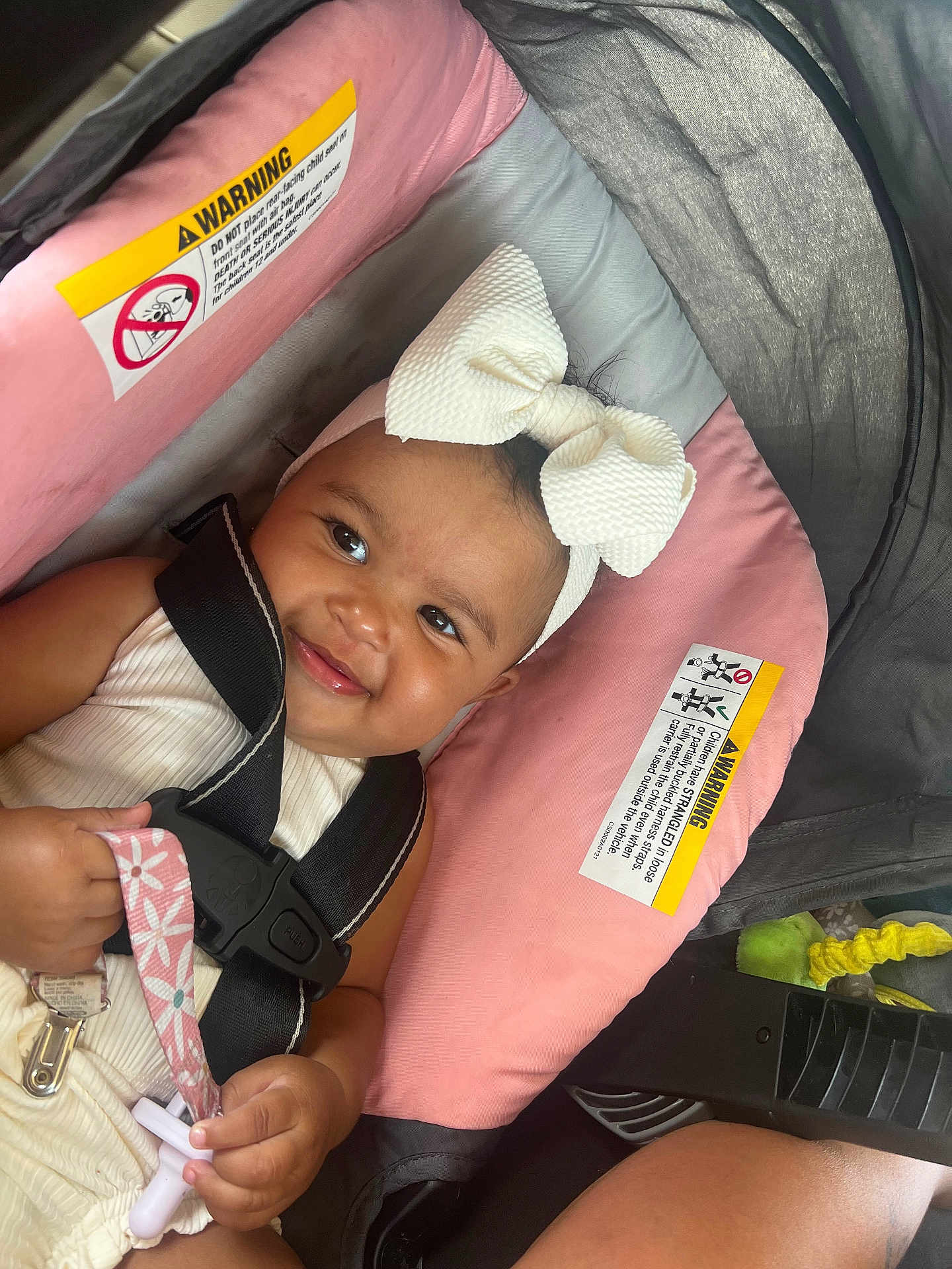 Ava is registered to the contest to win money with this photo: baby, child, smiling, headband, bow, car_seat, safety_harness, pink, gray, pacifier, hand, clothing, cute, infant, seat, happy, person, indoor, closeup, transportation