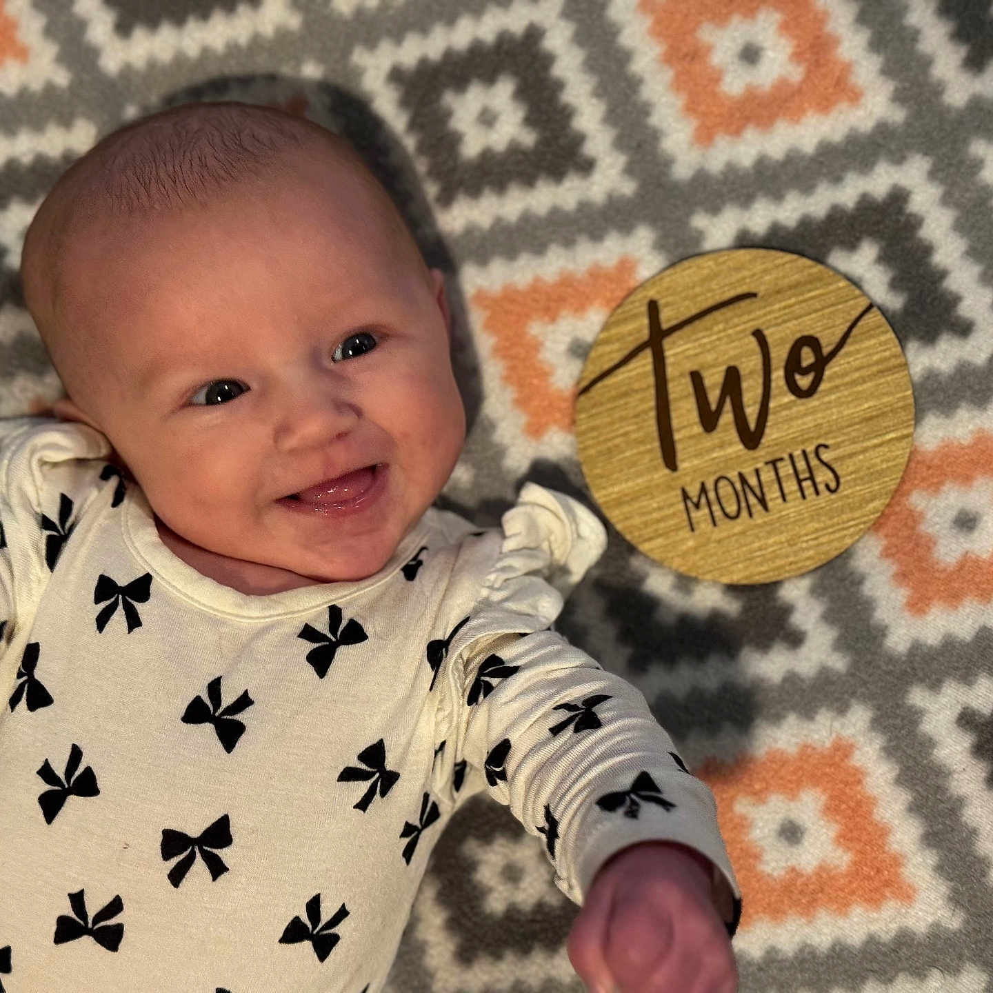Serena joined the competition — help win amazing prizes! arm, baby, black_bow_pattern, cheerful, child, cute, eyes, face, happy, head, infant, lying_down, mouth_open, onesie, patterned_blanket, skin, smiling, two_months, wooden_marker, young_baby