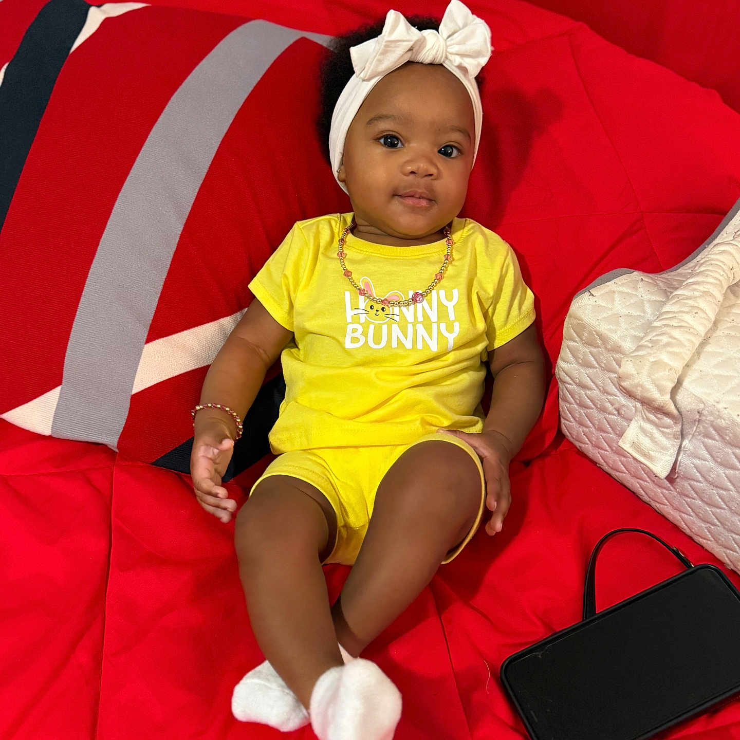 Blessyn is registered to the contest to win money with this photo: baby, child, yellow_clothing, headband, red_couch, pillow, quilted_bag, smartphone, sitting, indoors, cute, portrait, accessories, bracelet, white_socks, infant, happy, comfortable, modern, relaxed