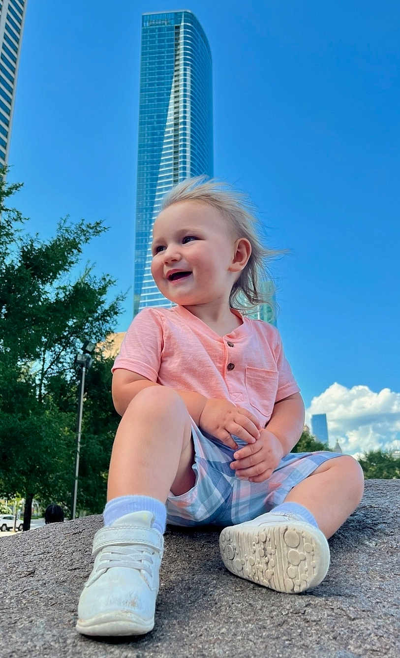 Joey joined the competition — help win amazing prizes! toddler, child, smiling, outdoor, urban, skyscraper, blue_sky, rock, casual_clothing, shorts, sneakers, trees, sunny, daytime, happy, portrait, nature, cityscape, person, playful