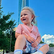Joseph is registered to the contest to win money with this photo: toddler, child, smiling, sitting, rock, urban, skyscraper, blue_sky, outdoor, casual_clothing, sneakers, shorts, shirt, happy, daytime, trees, nature, portrait, young_child, cityscape