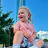 toddler, child, smiling, sitting, rock, urban, skyscraper, blue_sky, outdoor, casual_clothing, sneakers, shorts, shirt, happy, daytime, trees, nature, portrait, young_child, cityscape